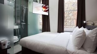 A hotel room containing a double bed with a shower unit at the end of the bed. There are two tall windows next to the bed.