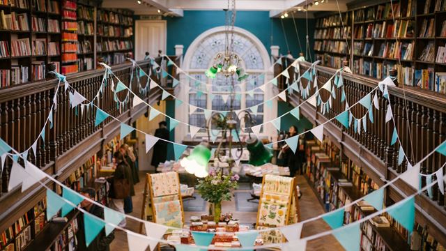 A view of Daunt Books with tall bookshelves, a central arched windows, and blue and pink bunting hanging from a wooden mezzanine area.