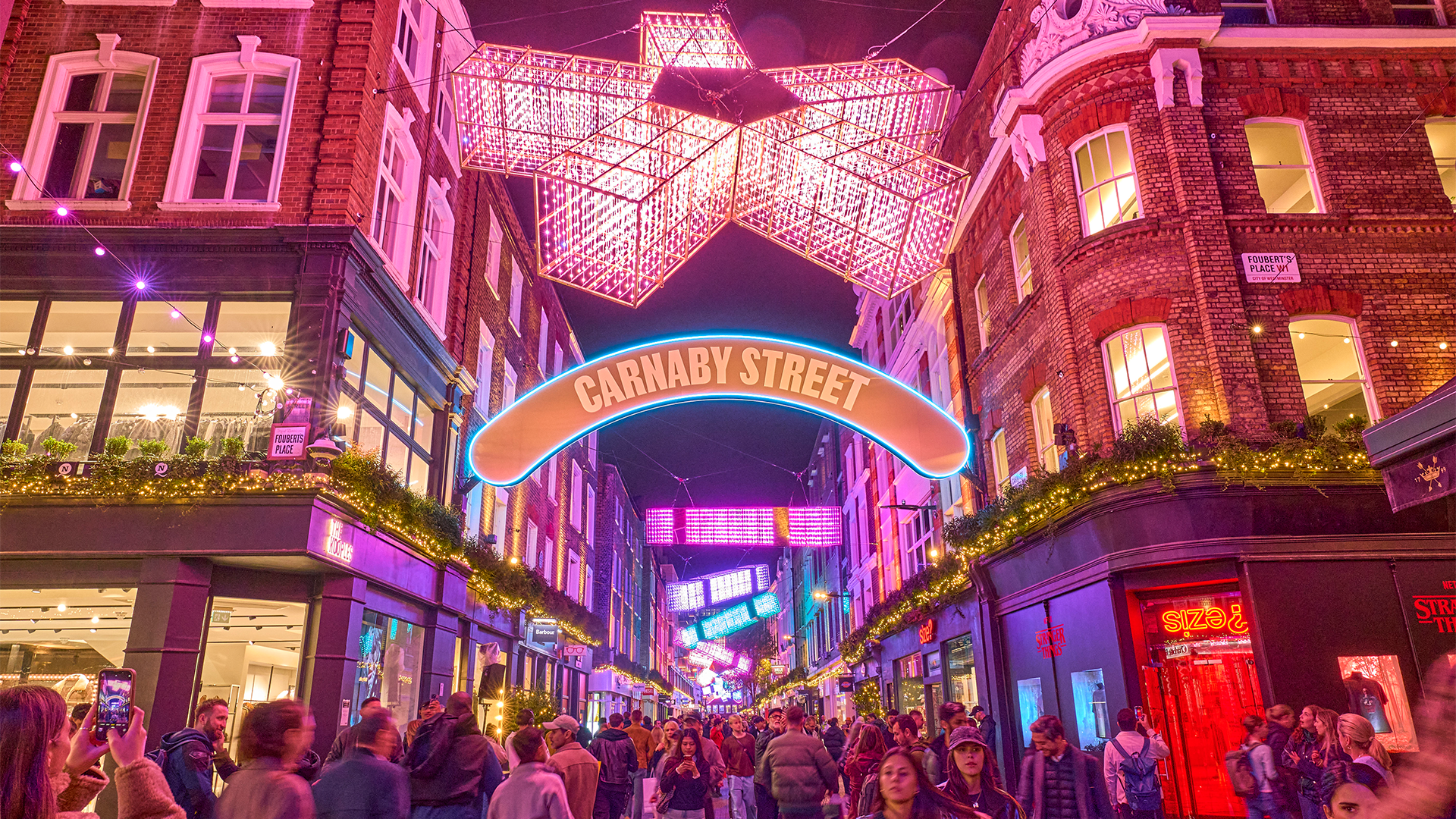 Admire the breathtaking festive display lining the famous Carnaby Street during a shopping trip. Credit: Shaftesbury Capital. Image courtesy of Shaftesbury Capital. Neon light installations hang above shoppers on Carnaby Street