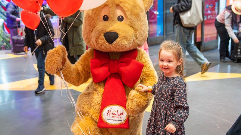 Hamleys is a magical place for kids at Christmas. Image courtesy of Hamleys. Child standing next to Hamleys bear figure holding balloons.