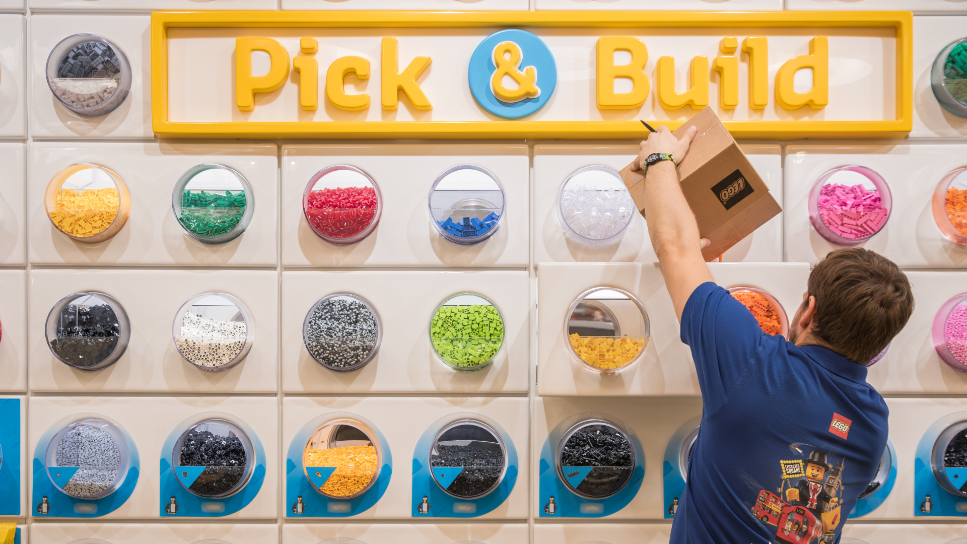 A man with a Lego shop uniform empties and box full of lego into one of the bubbles on the lego pick and build wall with lego pieces of every colour