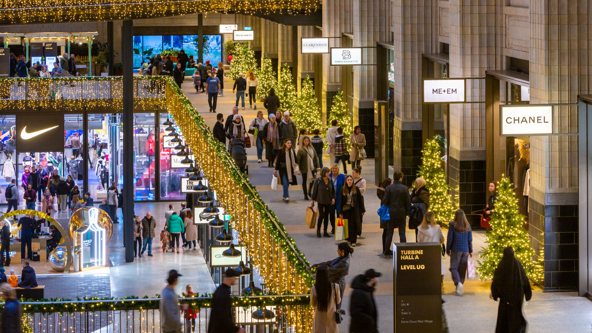 People walk around one of the many floors of shops inside Battersea Power Station adorned with Christmas decorations, with shop signs including Arcteryx, Reformation, SpaceNK and more lit up.