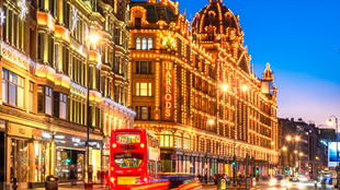 Warm twinkling lights cover the exterior of Harrods during Christmas during blue hour as light trails from cars and a red London bus sweep across the foreground