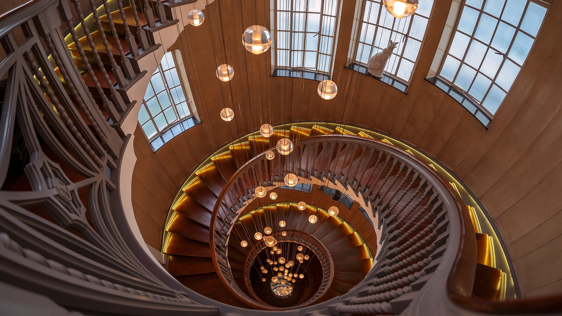 A view of the winding Heal's staircase looking down with its large windows and warm orb light pendants hanging in the middle of the staircase