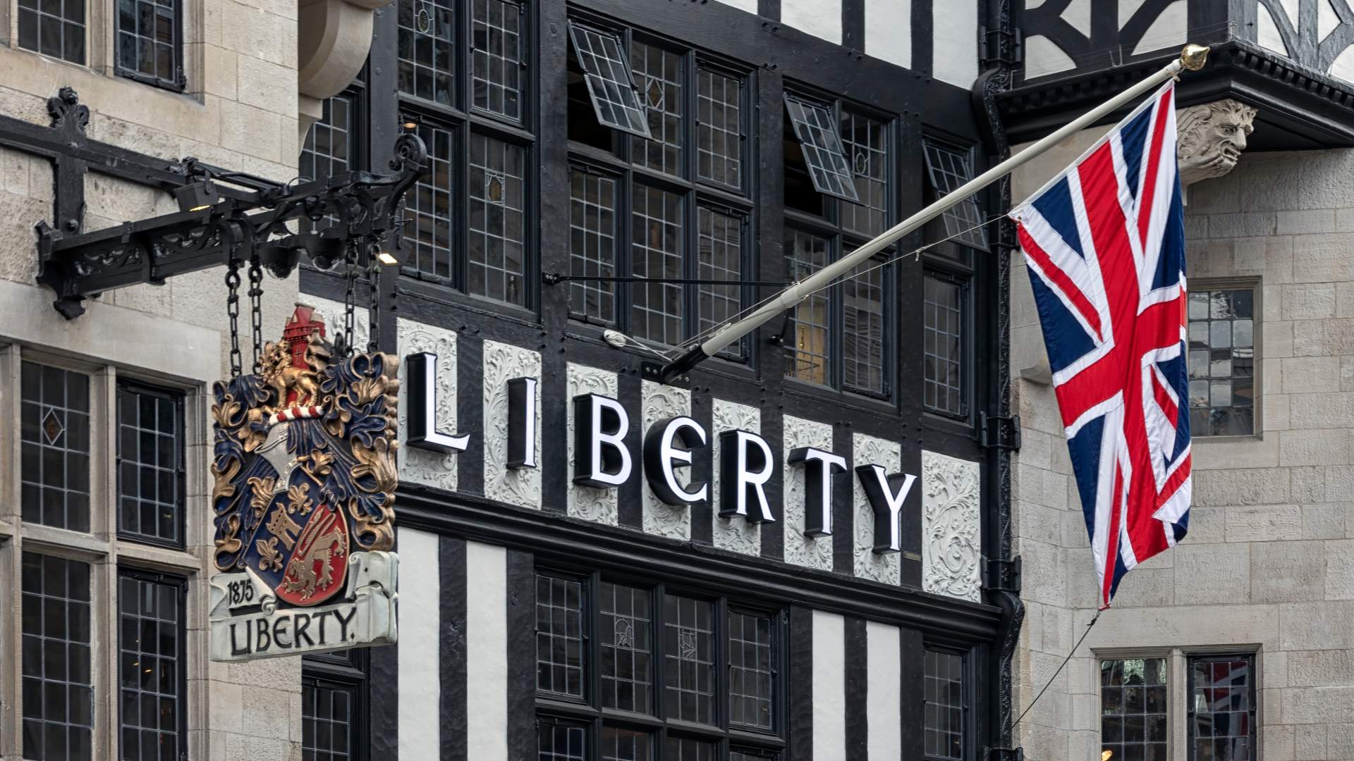A close up photo of the Liberty London sign with a Union Jack flag hanging above and a logo plaque hanging to the side against the iconic wooden beam exterior