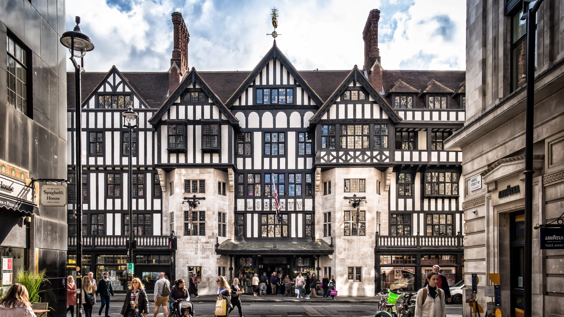 The front exterior of Liberty London in Soho with its iconic wooden beams, many windows and a busy street of pedestrians walking along the streets outside