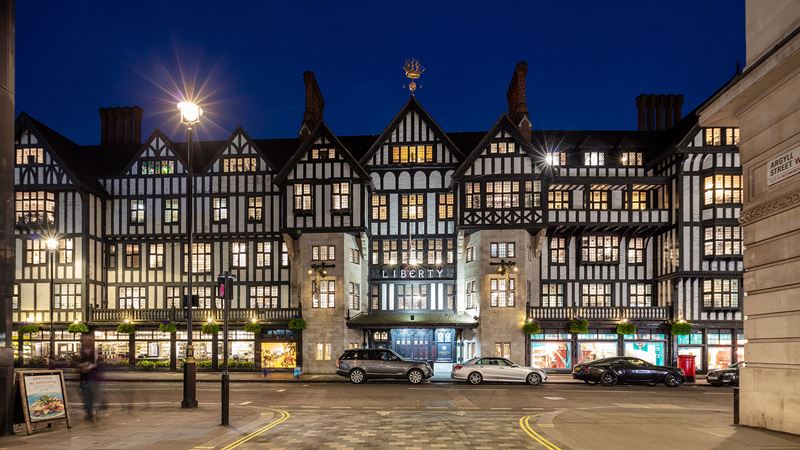 The Tudor-style black and white exterior of Libery London at night, with the sign "Liberty" above the door, and its windows illuminated from inside.