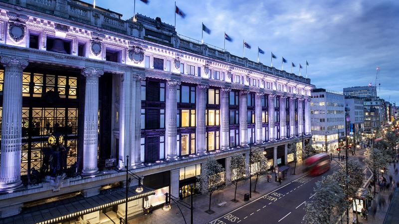 No London shopping trip is complete without a visit to Selfridges. Credit: Andrew Meredith. Image courtesy of Selfridges. A panoramic view from high of the facade of Selfridges, lit up in purpley-white lights, with Oxford Street below.
