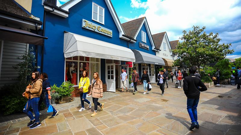 Enjoy luxury outlet shopping at Bicester Village, with designer brands, cafes and a stylish open‑air setting that is easy to reach by train from London. Image courtesy of Shutterstock. People walking past a row of colourful shops on a sunny day