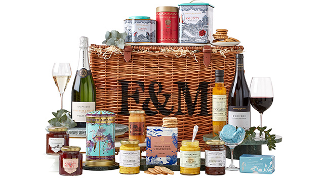 Un panier en osier avec le logo "F&M" écrit en noir, avec divers pots de confitures, de cornichons et de condiments, plus des bouteilles de vin et de vin mousseux, des boîtes de biscuits et d'autres aliments sur et autour du panier.