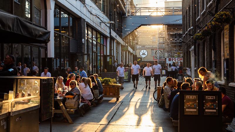 Browse, shop and eat your way around Brick Lane. Credit: Jon Reid. Image courtesy of visitlondon.com. Looking down a street in Spitalfields with people sitting outside pubs and restaurants each side and people walking towards the camera.