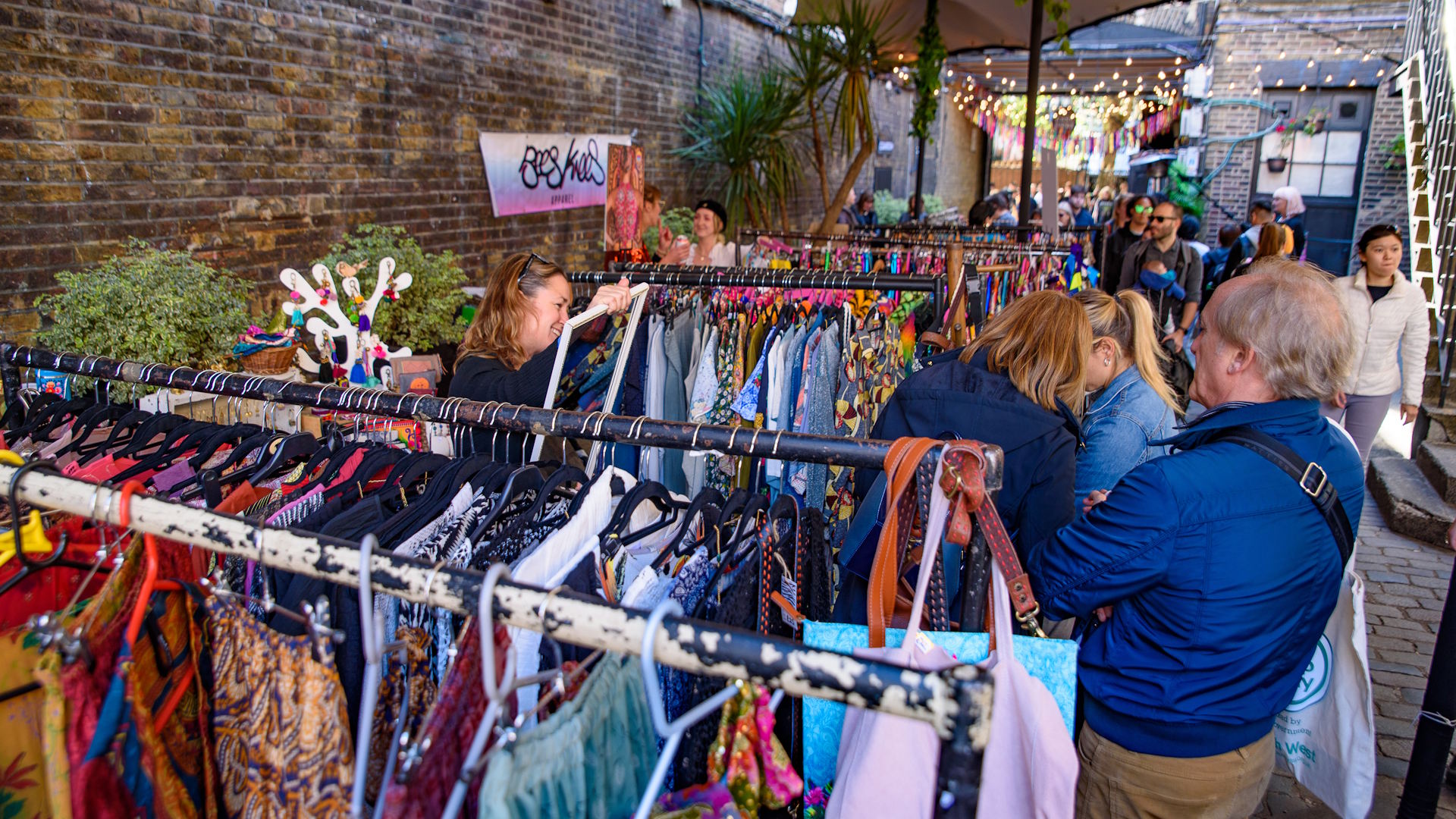 People browsing stalls seling vintage clothes outside with red-brick wall in the background.