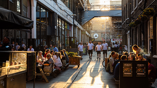 Looking down a street in Spitalfields with people sitting outside pubs and restaurants each side and people walking towards the camera.