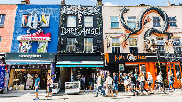 A terrace of colourful shops, painted in blue, black and cream, with large-scale sculptures of trainers and a dragon on front of the buildings. People walking past below, on a sunny day.