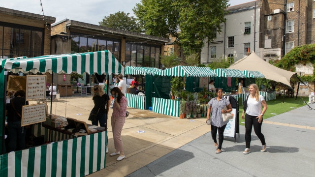 two women walking side by side conversing next to green and white striped market stalls 