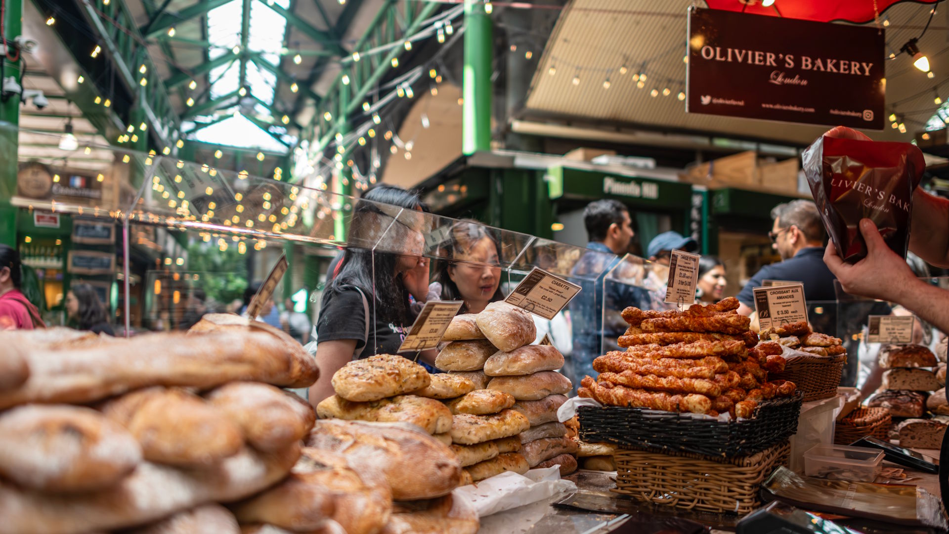 People being served at a bakery stall full of bread and baked goods within Borough Market, with the famous green struts of the market in the background.