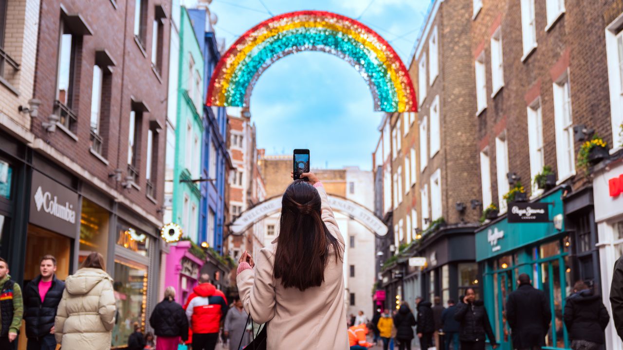 Shop until you drop on London's lively Carnaby Street. Photo credit: London & Partners. A woman in a white coat takes a photo of a rainbow decoration on Carnaby street in London