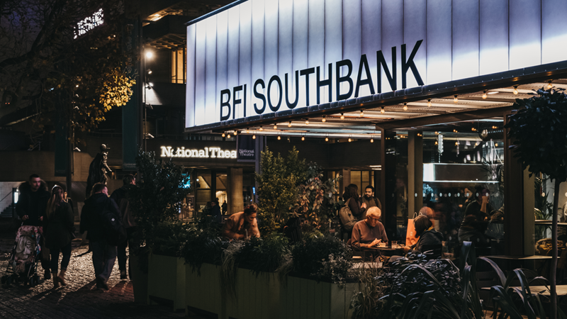 Celebrate the best new cinema at BFI London Film Festival in October. Credit: Shutterstock. Image courtesy of Shutterstock. An image of the front of BFI Southbank at night with groups of people sat outside at tables and walking in front of the building with lights illuminating the venue