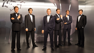 Six James Bond wax figures all stand together against a chrome background at Madame Tussauds, each wearing a black suit