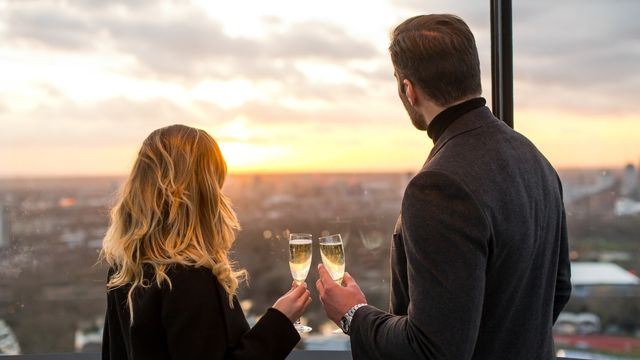 A couple stand looking out at a view of london holding glasses of champagne. 