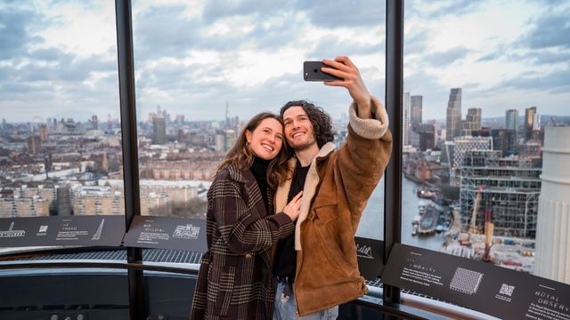 A couple stand close together as they take a selfie on battersea power station with London as the backdrop. 