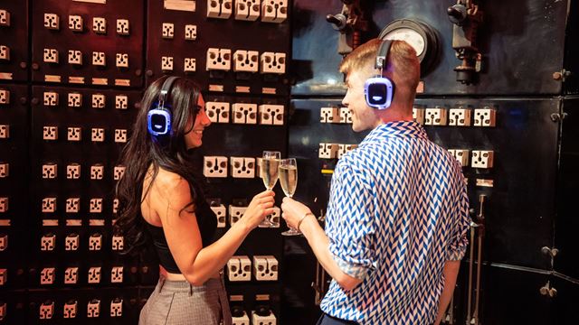 Two people wearing silent-disco headphones and holding champagne inside Control Room B at Battersea Power Station.