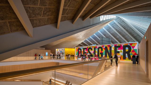 A large billboard with colourful triangles and the word "Designer" emblazoned across it, on the top floor of the Design Museum, with people walking past it.