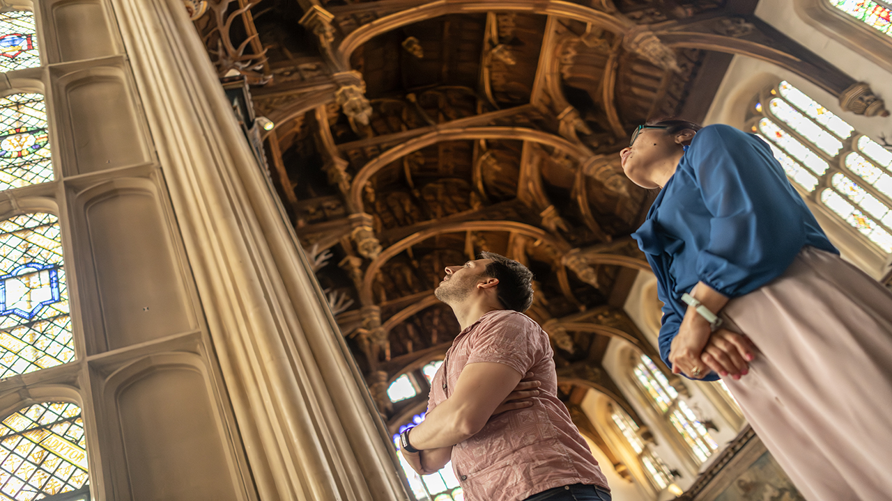 A man and a woman stand in one of the great halls of Hampton Court Palace and look up to admire the carved ceilings and stained glass windows.