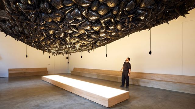 A man looks up at an art installation made from a large net holding what resemble large, shiny pebbles.