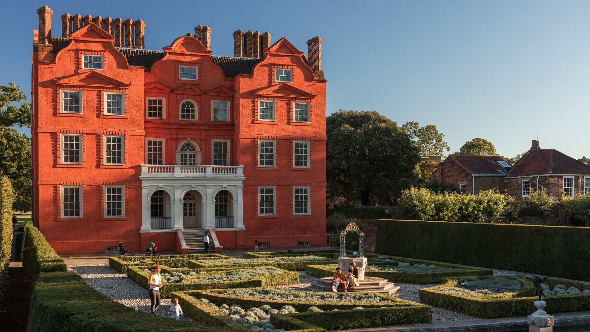 Kew Palace, a red brick royal residence, in the sunshine, with manicured hedges in front.