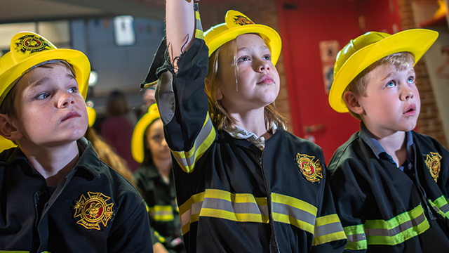 Une fille et deux garçons portent l'uniforme et le casque des pompiers et semblent prêts à partir avec leur équipe.