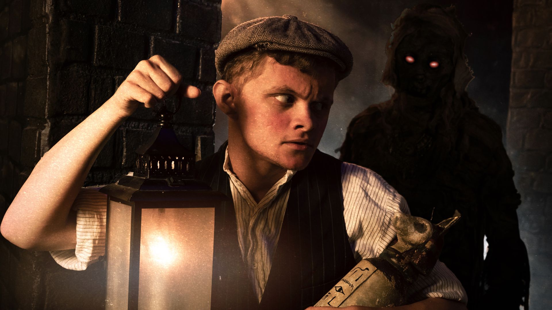A young man dressed in a white shirt, brown waistcoat and a brown flat cap holds a lantern and a small sarcophagus while a shadowy figure with red eyes hovers behind him. 