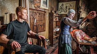 theatre set of a medieval doctors, with a visitor sat in a shackled chair and an actor pulling out a mannequins insides