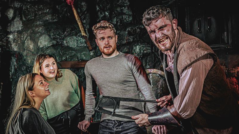 Visit London Dungeon's Torture Chamber to discover the medieval torture devices used on prisoners. Credit: The London Dungeon. Image courtesy of The London Dungeon. a visitor sat on the torture chair looking nervous, with two people watching smiling and a torture chamber actor holding a tool