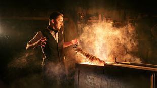 A performer stands beside a smoky display featuring a mummified skeleton in a dark, atmospheric setting.