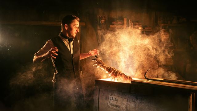 A performer stands beside a smoky display featuring a mummified skeleton in a dark, atmospheric setting.