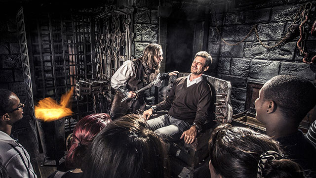 Der Foltermeister im The London Dungeon © The London Dungeon. Bild mit Genehmigung von The London Dungeon. Ein verängstigter Mann sitzt in einem Stuhl während ein andere Mann in einem mittelalterlichen Kostüm in foltert