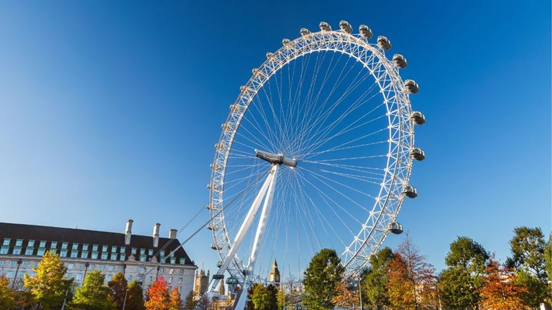 A woman walks along a path passing autumnal trees with clear skies and the London Eye in the background. 