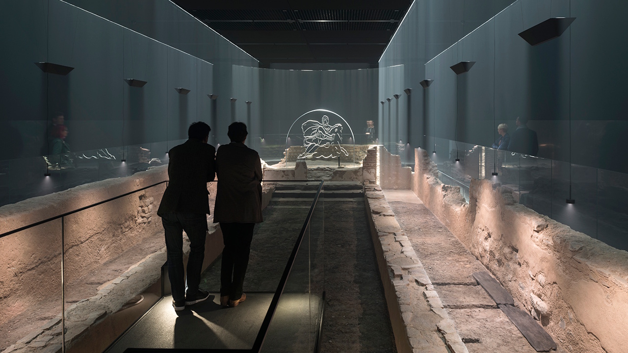 Two people look over the ruins of the Temple of Mithras, at the dimly lit London Mithraeum.