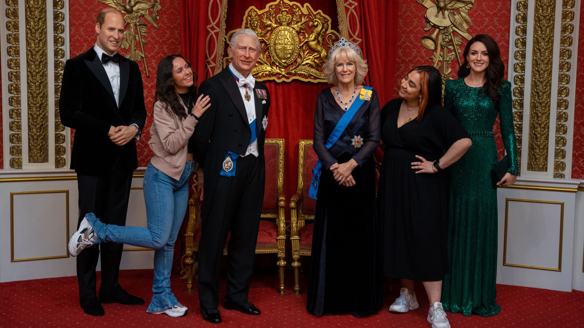 Pose with royalty at Madame Tussauds London. Image courtesy of Madame Tussauds London. three wax model figures of King Charles, Queen Camilla and Prince William in a red and gold decorated grand room, with two women smiling and posing for a photo