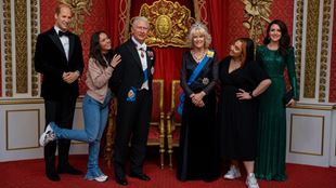 three wax model figures of King Charles, Queen Camilla and Prince William in a red and gold decorated grand room, with two women smiling and posing for a photo