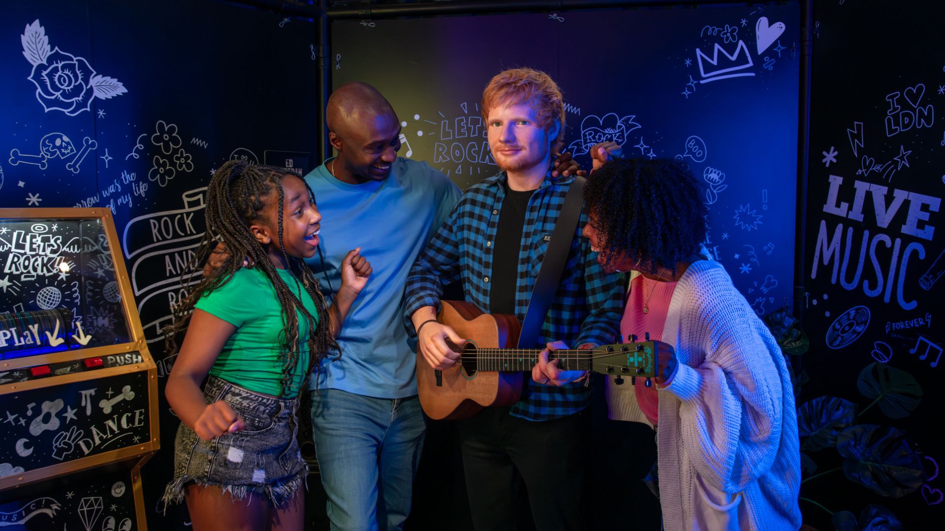 Meet iconic British singer-songwriter Ed Sheeran and pose in the live music space. Image courtesy of Madame Tussauds London. wax figure model of Ed Sheeran, wearing a black tshirt, jeans and a green chequered shirt over the top, smiling and holding a guitar. three people stand around the model smiling