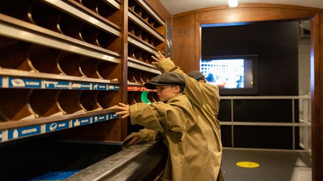 two young children dressed up in oversized mail worker outfits, pretending to file post into the cupboards