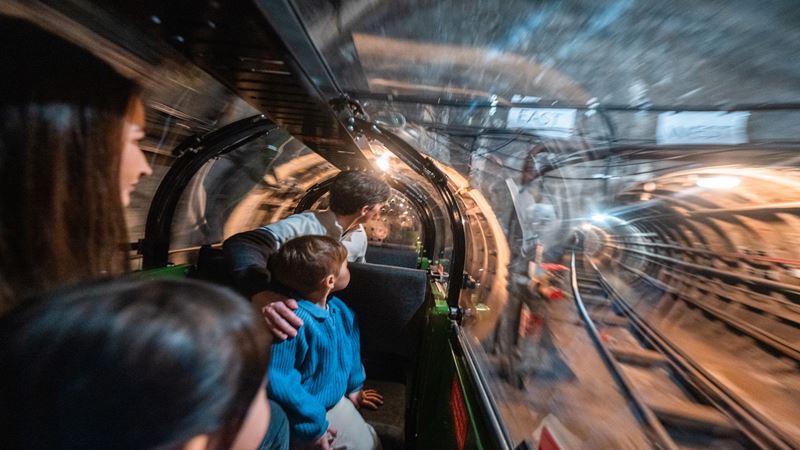 Ride the Mail Rail at The Postal Museum. Image courtesy of The Postal Museum. a family of four sat inside a narrow train with a glass roof, looking out to see the train track tunnel ahead