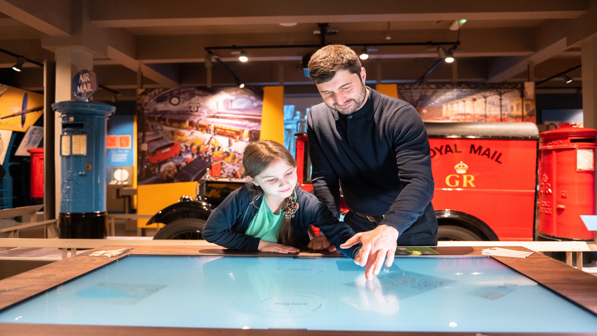 Erkunden Sie interaktive Ausstellungen und erfahren Sie mehr über die faszinierende Geschichte der Post. Bild mit Genehmigung von The Postal Museum. Ein Mann und ein Kind betrachten einen interaktiven Ausstellungstisch mit Postmemorabilien im Hintergrund