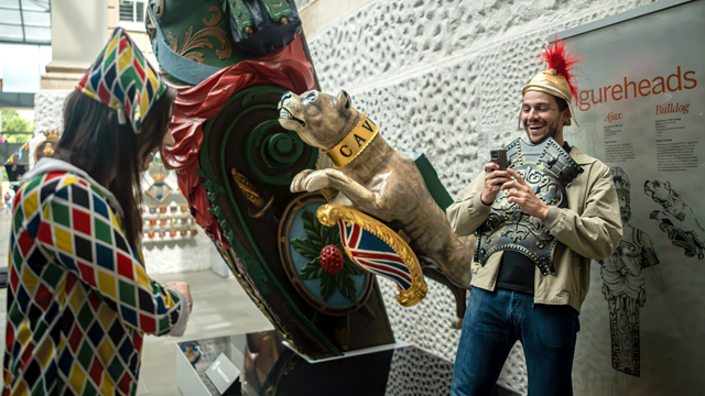 Un homme portant un casque de soldat romain sourit en prenant une photo d'une femme habillée d'une tenue d'arlequin au National Maritime Museum.