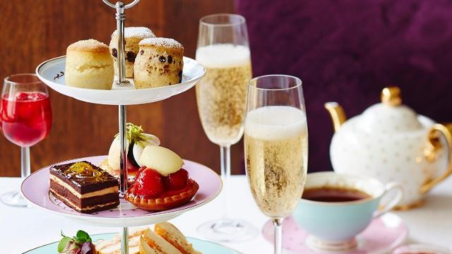 A table covered in sweet treaats including scones, chocolate cake and strawberry deserts with a white and gold teapot and two glasses of bubbly champagne.