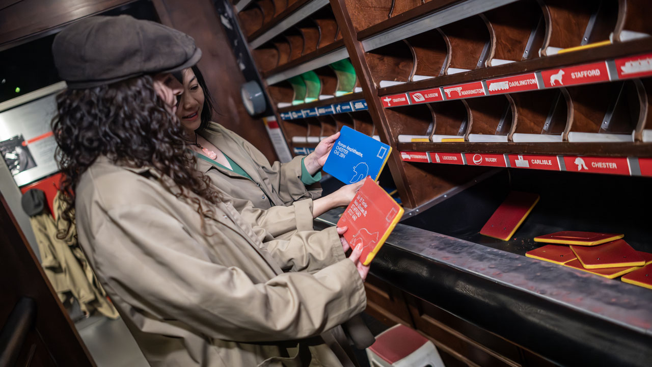 Visitez le Postal Museum, une attraction immersive explorant le système postal londonien et ses meilleures inventions. ©London & Partners/Michael Barrow Deux femmes, vêtues de trenchs et de casquettes grises, trient le courrier au Musée de la Poste.