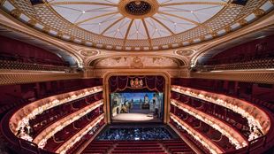 A view of the Royal Opera House auditorium with its red velvet seats beneath a golden ceiling.