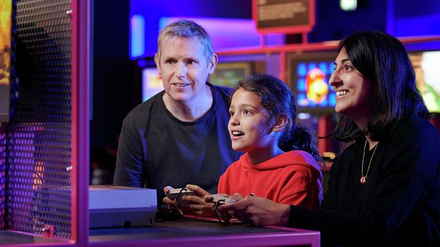 Family crowded round games station with child holding games console.
